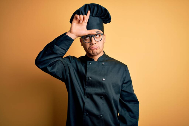 Mladý brazilský kuchař muž na sobě vařič uniformu a klobouk přes izolované žluté pozadí dělat si legraci z lidí s prsty na čele dělat poražený gesto posměch a urážlivé. - Fotografie, Obrázek