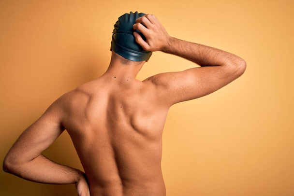 Mladý pohledný muž bez trička na sobě plavky a plavky čepice přes izolované žluté pozadí pozpátku přemýšlet o pochybách s rukou na hlavě - Fotografie, Obrázek