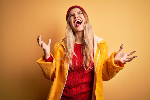 Young beautiful blonde woman wearing raincoat and wool cap over isolated yellow background crazy and mad shouting and yelling with aggressive expression and arms raised. Frustration concept. - Photo, Image