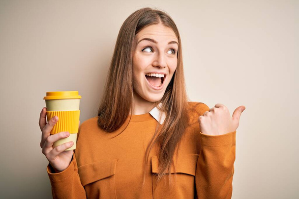 若いです美しいです赤毛女性飲むカップのコーヒー隔離された白い背景ポインティングと見せますとともに親指で側まで幸せな顔笑顔 - 写真・画像
