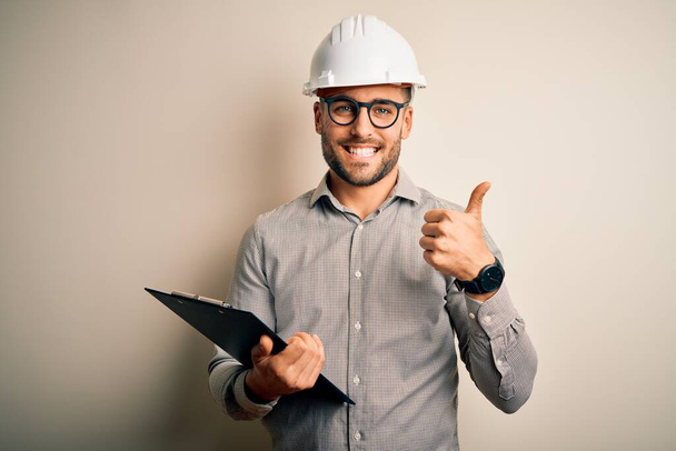 Młody architekt inspektor człowiek noszący kask bezpieczeństwa budowniczy i za pomocą schowka szczęśliwy z dużym uśmiechem robi znak ok, kciuk w górę palcami, doskonały znak - Zdjęcie, obraz