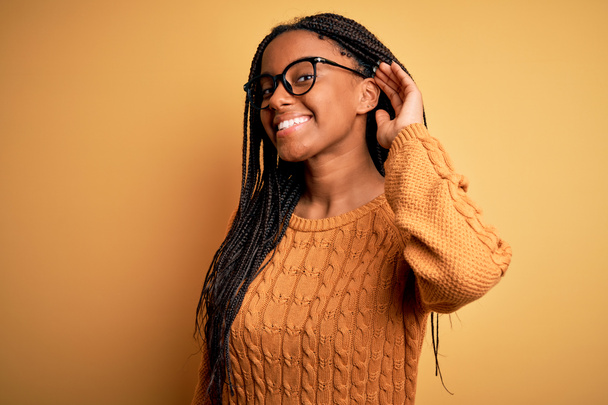 Νεαρή Αφροαμερικανή έξυπνη γυναίκα που φοράει γυαλιά και casual πουλόβερ πάνω από κίτρινο φόντο χαμογελώντας με το χέρι πάνω από το αυτί ακούγοντας μια ακρόαση για φήμες ή κουτσομπολιά. Έννοια κώφωση. - Φωτογραφία, εικόνα