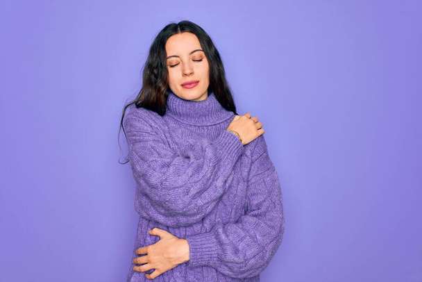 Young beautiful woman wearing casual turtleneck sweater standing over purple background Hugging oneself happy and positive, smiling confident. Self love and self care - Photo, Image