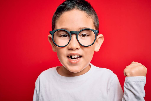 Νεαρό μικρό έξυπνο παιδί φορώντας γυαλιά nerd πάνω από το κόκκινο απομονωμένο φόντο ουρλιάζοντας υπερήφανος και γιορτάζει τη νίκη και την επιτυχία πολύ ενθουσιασμένος, ζητωκραυγάζοντας συναίσθημα - Φωτογραφία, εικόνα