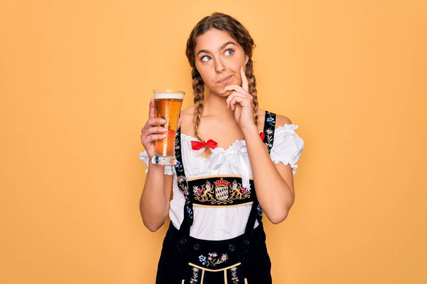 Mooie blonde Duitse vrouw met blauwe ogen dragen octoberfest jurk drinken glas bier serieus gezicht denken over vraag, zeer verward idee - Foto, afbeelding