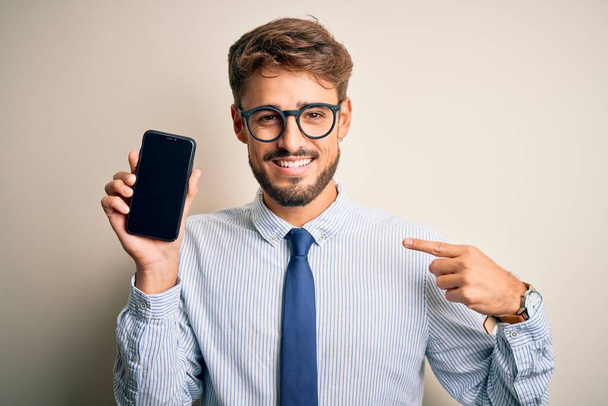 Молодий бізнесмен тримає смартфон, що стоїть над ізольованим білим тлом з несподіваним обличчям, спрямованим пальцем до себе
 - Фото, зображення