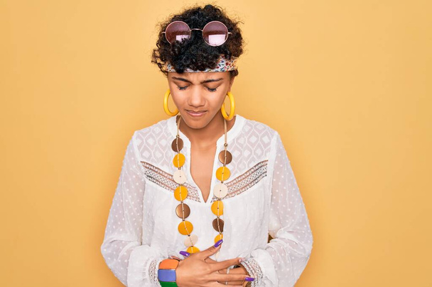 Jonge mooie Afrikaanse Amerikaanse afro hippie vrouw draagt een zonnebril en accessoires met de hand op de maag omdat indigestie, pijnlijke ziekte zich onwel voelt. Ache-concept. - Foto, afbeelding