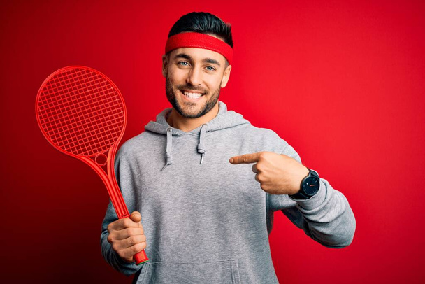 Jeune beau sportif tenant une raquette de tennis portant des vêtements de sport sur fond rouge avec un visage surprise pointant du doigt vers lui-même
 - Photo, image