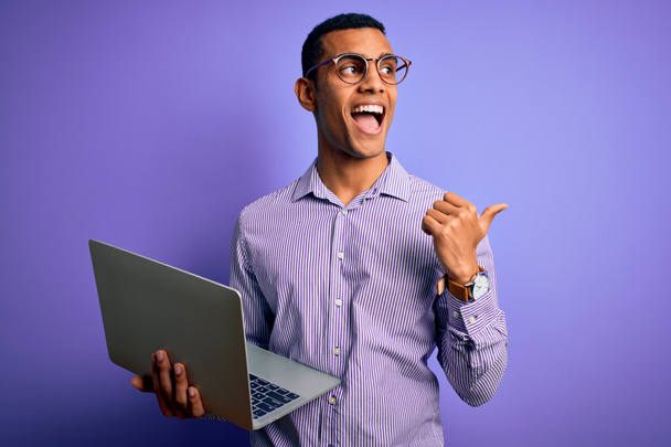 Genç ve yakışıklı Afro-Amerikan iş adamı laptopunu mor arka planda kullanıyor ve gülümseyen bir gülümsemeyle baş parmağıyla gösteriyor. - Fotoğraf, Görsel
