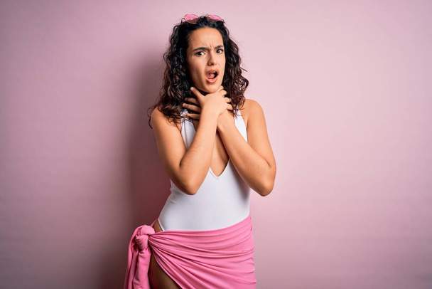 Beautiful woman with curly hair on vacation wearing white swimsuit over pink background shouting suffocate because painful strangle. Health problem. Asphyxiate and suicide concept. - Photo, Image