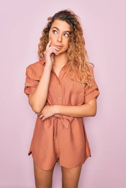 Young beautiful woman with blue eyes wearing casual summer dress over pink background with hand on chin thinking about question, pensive expression. Smiling with thoughtful face. Doubt concept. - Fotó, kép