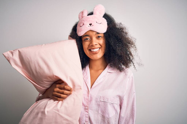 Young african american woman with afro hair wearing pajama and sleep mask holding pillow with a happy face standing and smiling with a confident smile showing teeth - Photo, Image