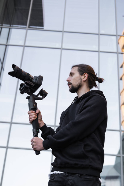 Young Professional videographer holding professional camera on 3-axis gimbal stabilizer. Pro equipment helps to make high quality video without shaking. Cameraman wearing white hoodie making a videos. - Fotoğraf, Görsel
