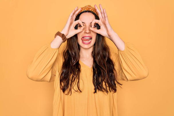 Young beautiful woman wearing casual t-shirt and diadem standing over yellow background doing ok gesture like binoculars sticking tongue out, eyes looking through fingers. Crazy expression. - Photo, Image