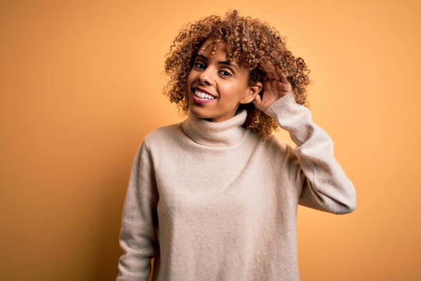 Fiatal, gyönyörű afro-amerikai nő garbó pulóvert visel sárga háttér felett mosolyogva, fültől fülig hallgatva egy pletykát vagy pletykát. Süketség fogalma. - Fotó, kép