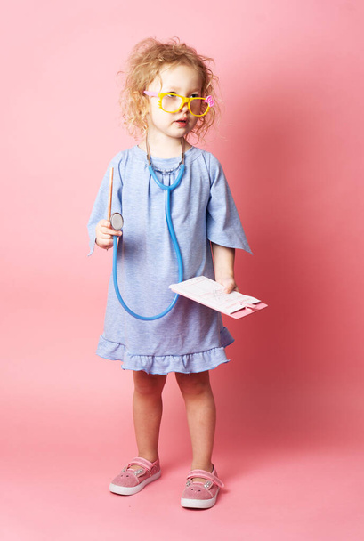 Pediatrics: a little girl in the form of a nurse with glasses and with a stethoscope fills a prescription. Portrait on a pink background. - Photo, Image