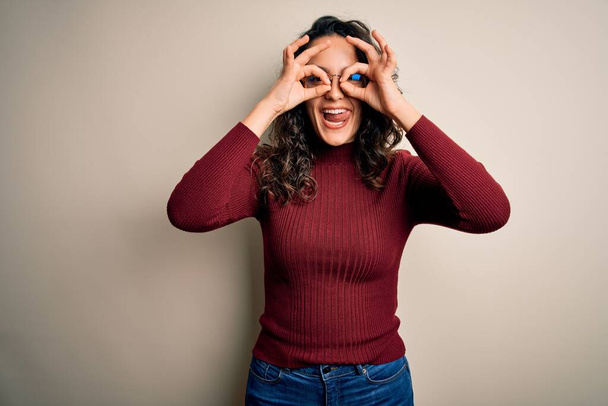 Beautiful woman with curly hair wearing casual sweater and glasses over white background doing ok gesture like binoculars sticking tongue out, eyes looking through fingers. Crazy expression. - Фото, зображення