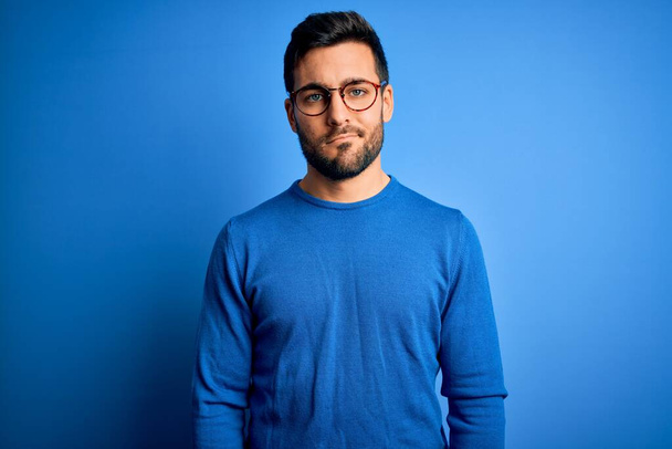 Young handsome man with beard wearing casual sweater and glasses over blue background looking sleepy and tired, exhausted for fatigue and hangover, lazy eyes in the morning. - Фото, изображение