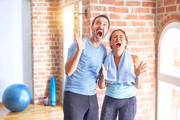 Middle age sporty couple standing before do exercise at gym crazy and mad shouting and yelling with aggressive expression and arms raised. Frustration concept. - Photo, Image