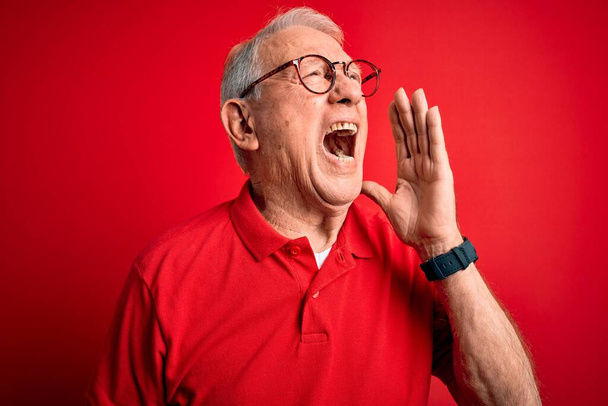 赤い背景に眼鏡とカジュアルなTシャツを着た白髪の先輩が大声で叫び、口の中で手で横に大声で叫んでいます。コミュニケーションの概念. - 写真・画像