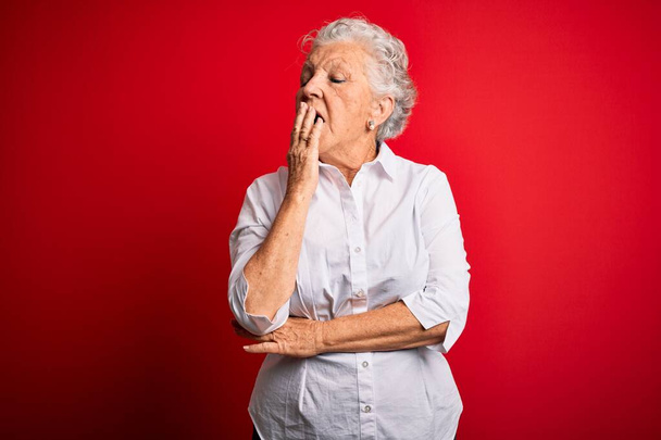 Старшая красивая женщина в элегантной рубашке, стоящей на изолированном красном фоне скучает зевая усталый закрывающий рот рукой. Беспокойство и сонливость
. - Фото, изображение
