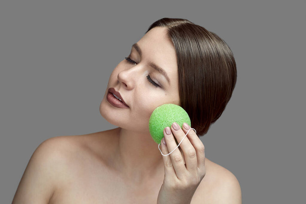 chica hacer un tratamiento facial con una esponja verde konjac sobre un fondo gris con el lugar para tex
 - Foto, imagen