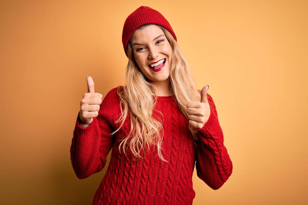 Young beautiful blonde woman wearing casual sweater and wool cap over white background success sign doing positive gesture with hand, thumbs up smiling and happy. Cheerful expression and winner gesture. - Photo, Image