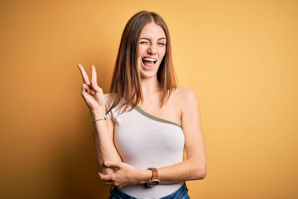 Молодая красивая рыжая женщина в повседневной футболке на изолированном желтом фоне улыбается со счастливым лицом, подмигивая в камеру, делая знак победы пальцами. Номер два:
. - Фото, изображение