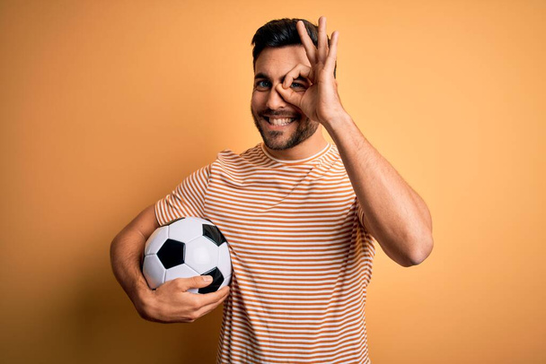 Hombre guapo jugador con barba jugando fútbol sosteniendo el balón de fútbol sobre fondo amarillo haciendo buen gesto con la mano sonriente, ojos mirando a través de los dedos con la cara feliz
. - Foto, imagen