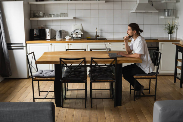 Freelancer working from home on the kitchen and using laptop. Bearded man working with a laptop and reading good news. Handsome successful self entrepreneur sitting and working at his modern home. - Foto, Bild