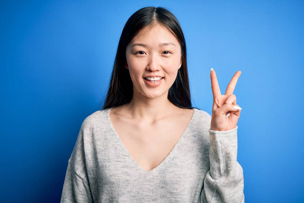 Fiatal, gyönyörű ázsiai nő, alkalmi pulóvert visel, kék, elszigetelt háttér felett áll, és a kettes számú ujjaival mutogat, miközben magabiztosan és boldogan mosolyog.. - Fotó, kép