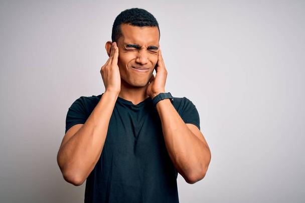 Jeune bel homme afro-américain portant un t-shirt décontracté debout sur fond blanc couvrant les oreilles avec des doigts avec une expression agacée pour le bruit de la musique forte. Concept sourd
. - Photo, image