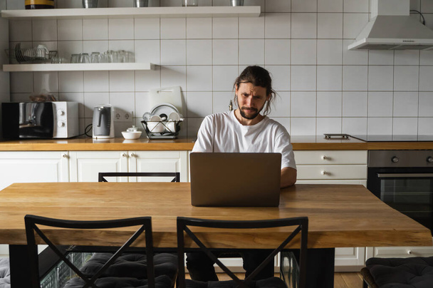 Freelancer working from home on the kitchen and using laptop. Bearded man working with a laptop and reading bad news. Handsome successful self entrepreneur sitting and working at his modern home. - Foto, Imagen