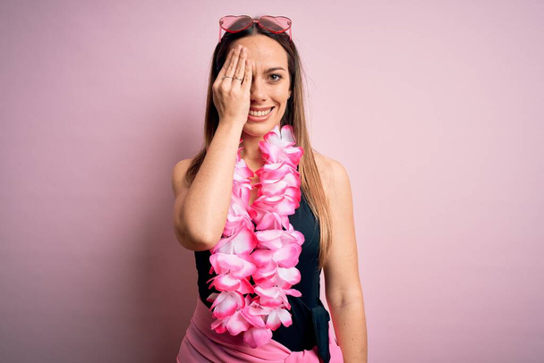 Νεαρή όμορφη ξανθιά γυναίκα με μαγιό και λουλουδάτο χαβανέζικο lei πάνω από ροζ φόντο που καλύπτει το ένα μάτι με το χέρι, αυτοπεποίθηση χαμόγελο στο πρόσωπο και συγκίνηση έκπληξη. - Φωτογραφία, εικόνα