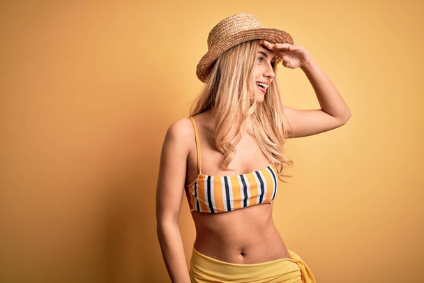 Young beautiful blonde woman on vacation wearing bikini and hat over yellow background very happy and smiling looking far away with hand over head. Searching concept. - Photo, image