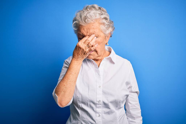 Starsza piękna kobieta ubrana w elegancką koszulę stojąca nad odizolowanym niebieskim tłem zmęczona pocieraniem nosa i oczu uczucie zmęczenia i bólu głowy. Koncepcja stresu i frustracji. - Zdjęcie, obraz