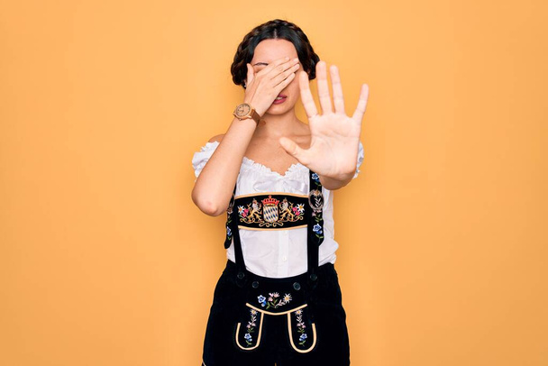 Young beautiful german woman with blue eyes wearing traditional octoberfest dress covering eyes with hands and doing stop gesture with sad and fear expression. Embarrassed and negative concept. - Photo, Image