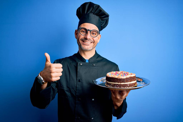 Joven panadero guapo con uniforme de cocina y sombrero sosteniendo bandeja con pastel feliz con gran sonrisa haciendo signo ok, pulgar hacia arriba con los dedos, señal excelente
 - Foto, imagen