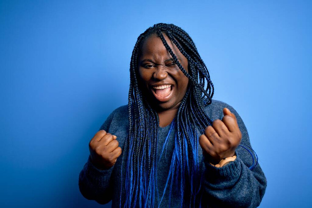 Femme afro-américaine plus la taille avec des tresses portant un pull décontracté sur fond bleu célébrant surpris et étonnés pour le succès avec les bras levés et les yeux fermés. Concept gagnant
. - Photo, image