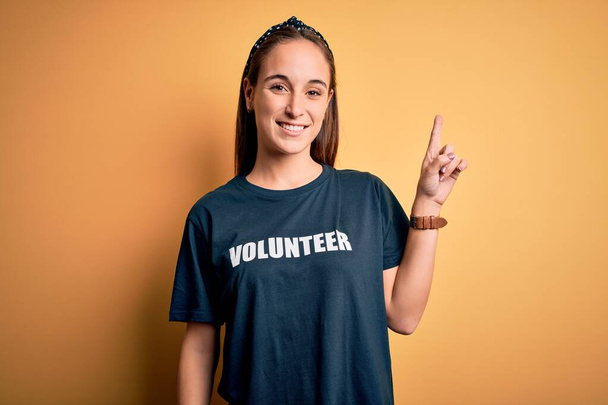 Młoda piękna kobieta w ochotniczej koszulce robi wolontariat na żółtym tle z dużym uśmiechem na twarzy, wskazując palcem w bok, patrząc w kamerę.. - Zdjęcie, obraz
