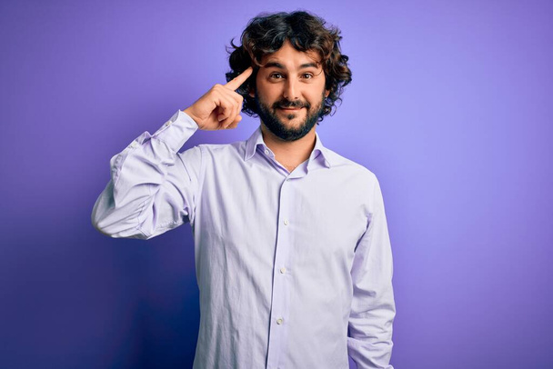 Mladý pohledný obchodník s vousy v košili stojící nad fialovým pozadím Usmívající se ukazující na hlavu jedním prstem, skvělý nápad nebo myšlenka, dobrá paměť - Fotografie, Obrázek