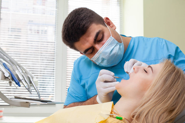 Dentista examinando los dientes del paciente, vistiendo uniforme azul y guantes, mirando los dientes del paciente cuidadosamente con instrumentos dentales. Hermosa mujer rubia visitando dentista
 - Foto, Imagen