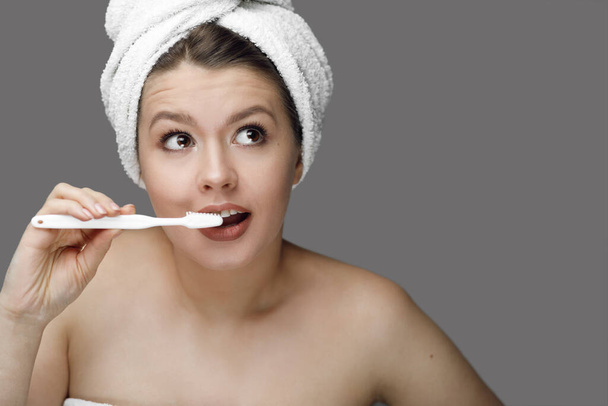 κορίτσι με μια λευκή οδοντόβουρτσα σε μια πετσέτα στο κεφάλι της σε ένα γκρι φόντο με το μέρος για tex - Φωτογραφία, εικόνα