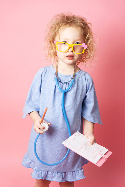 Pediatrics: a little girl in the form of a nurse with glasses and with a stethoscope fills a prescription. Portrait on a pink background. - Zdjęcie, obraz