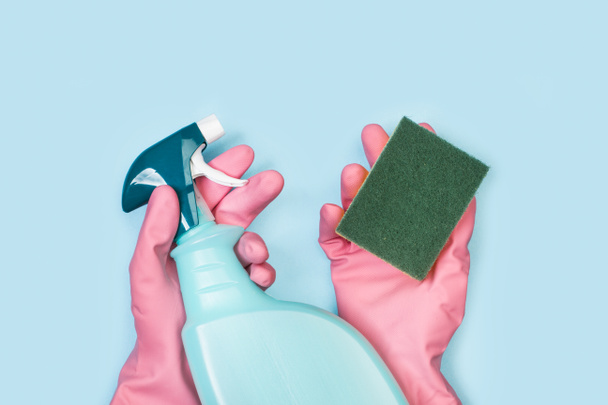 Mains avec des gants en caoutchouc rose tenant une éponge nettoyante et un pulvérisateur de produits nettoyants sur un fond bleu clair
 - Photo, image