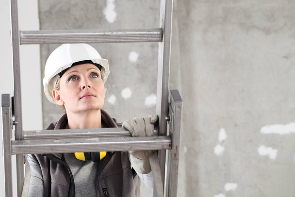 donna costruttore lavoratore edile su scala indossando casco bianco e cuffie di protezione dell'udito sul sito interno costruzione sfondo - Foto, immagini