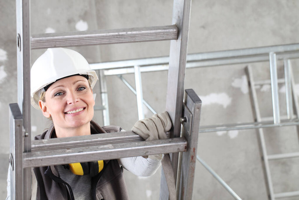 mujer sonriente constructora de trabajadores de la construcción en escalera con casco blanco y audífonos de protección auditiva en el fondo del edificio del sitio interior - Foto, imagen