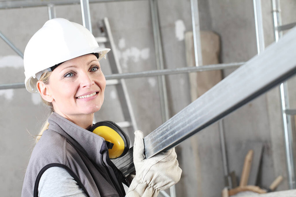 Lächelnde Bauarbeiterin Bauarbeiter-Porträt mit weißem Helm und Gehörschutz-Kopfhörer, hält einen Metallstift für Trockenbau auf der Baustelle im Inneren mit Gerüsten - Foto, Bild