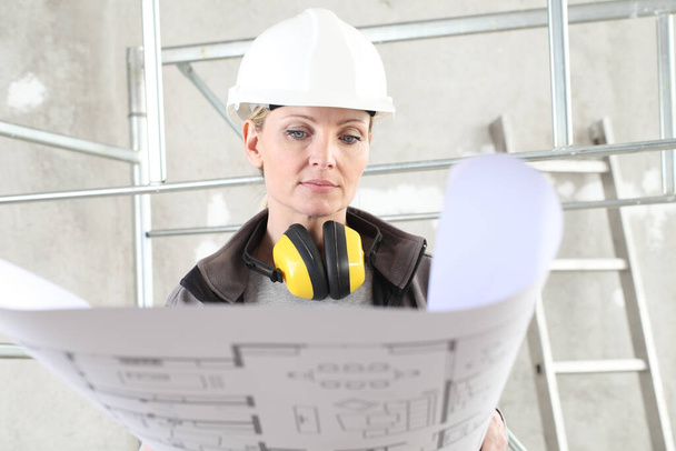 İnşaat işçisi kadın inşaatçı planlara bakıyor, miğfer takıyor ve bina arka planında kulaklık takıyor. - Fotoğraf, Görsel