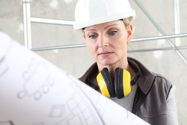 vrouw bouwvakker kijken naar blauwdruk, het dragen van een helm en gehoorbescherming koptelefoon in de bouwplaats binnenshuis achtergrond - Foto, afbeelding